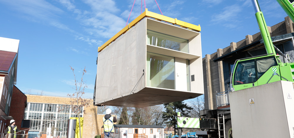 Image of a construction site, showing a portable building being moved into place by a green crane